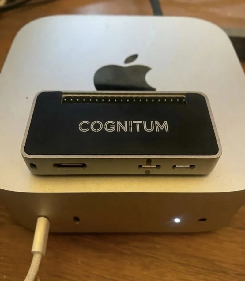 The Cognitum One Seed photographed front-facing — flat matte-black enclosure with the live LED matrix display visible across the front face, sitting atop a Mac mini.